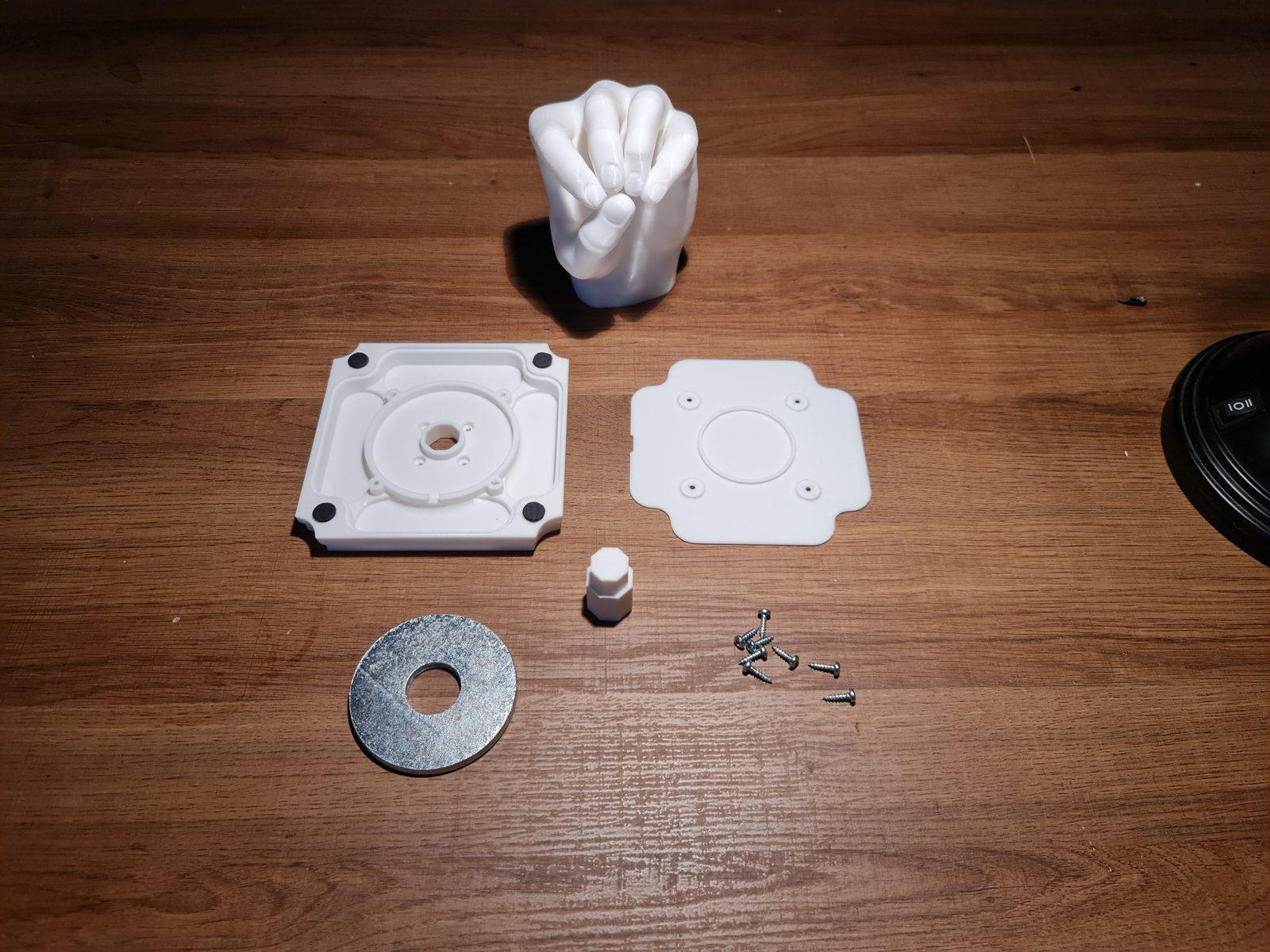 The parts of one piece laid out on a table: a fist hand, the base, the cover plate, the peg, a steel washer and screws